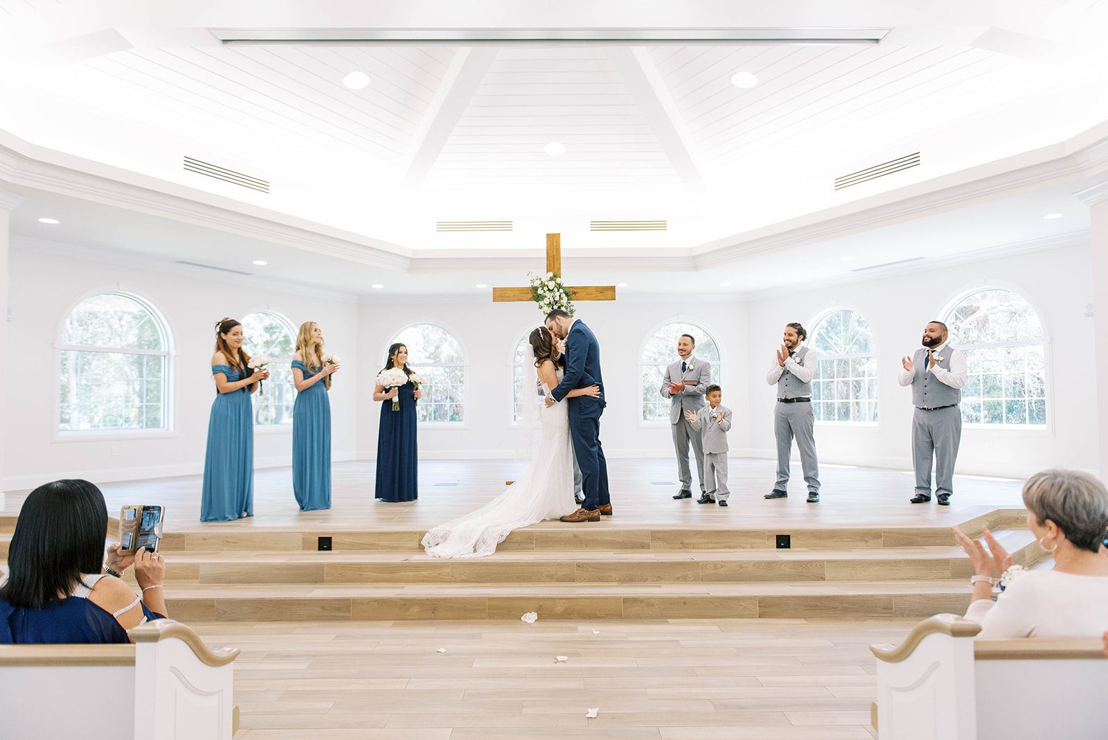 Harborside Chapel wedding with Puerto Rican couple kissing in front of a wooden cross with their wedding party to their sides 
