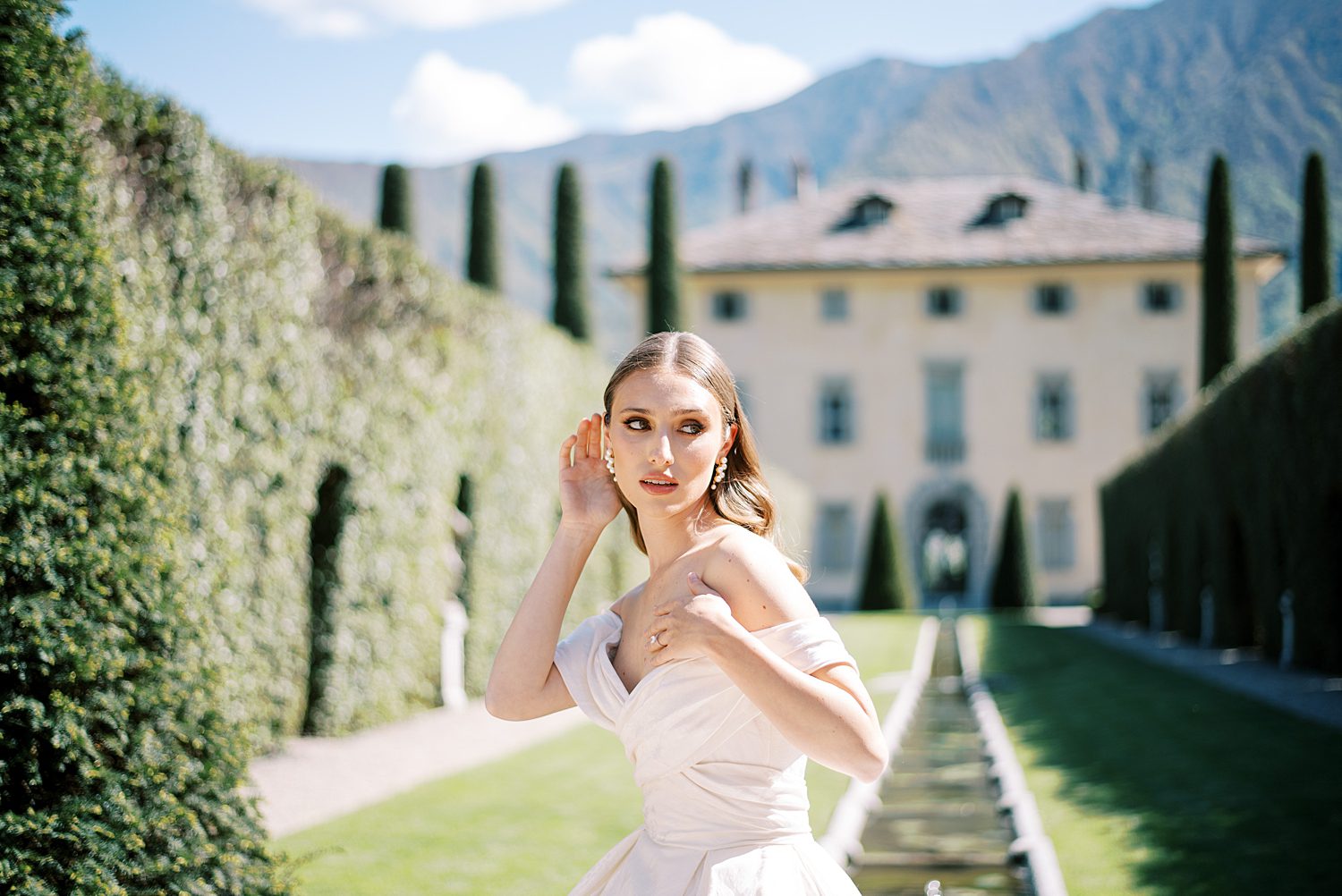brunette bride pushes hair behind ear in off-the-shoulder gown in front of Villa Balbiano