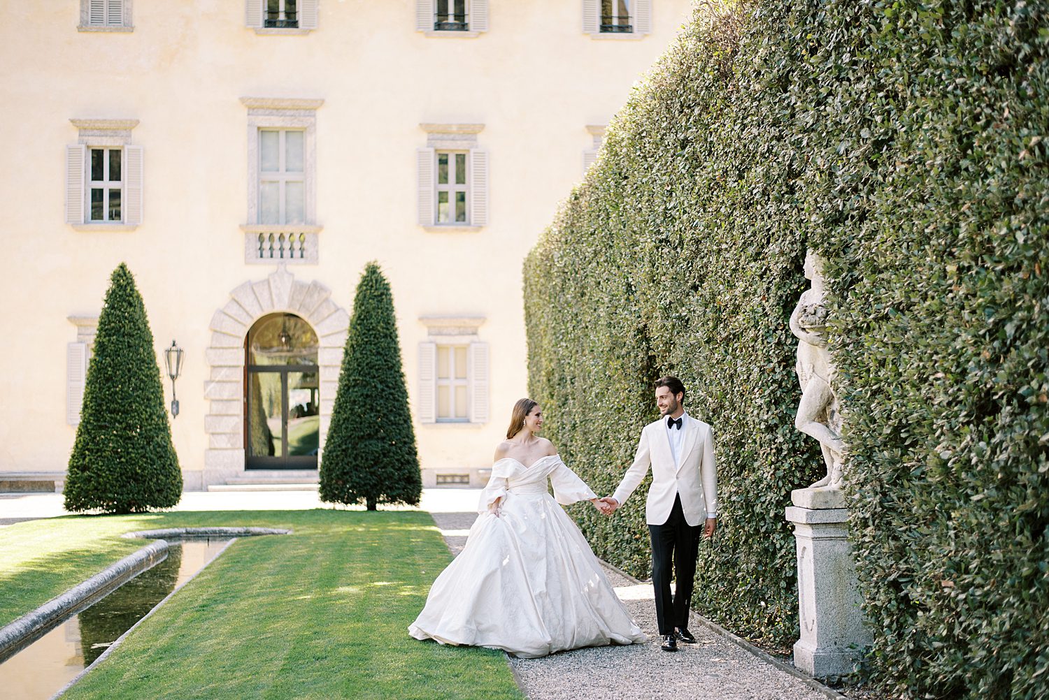 bride and groom hold hands walking along hedges in front of Villa Balbiano