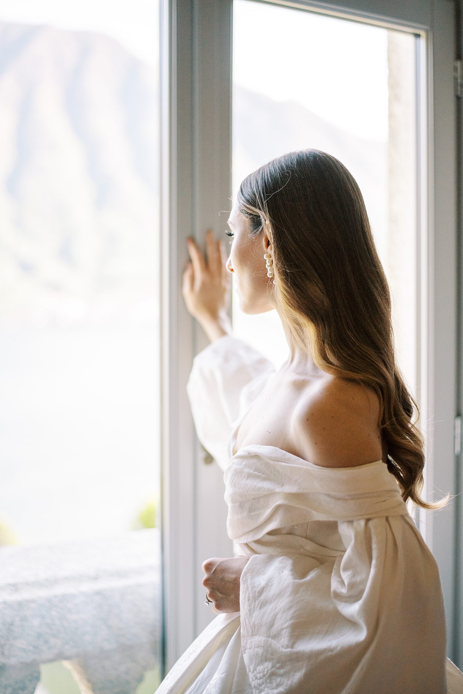 bride looks out window at Villa Balbiano with off-the-shoulder gown