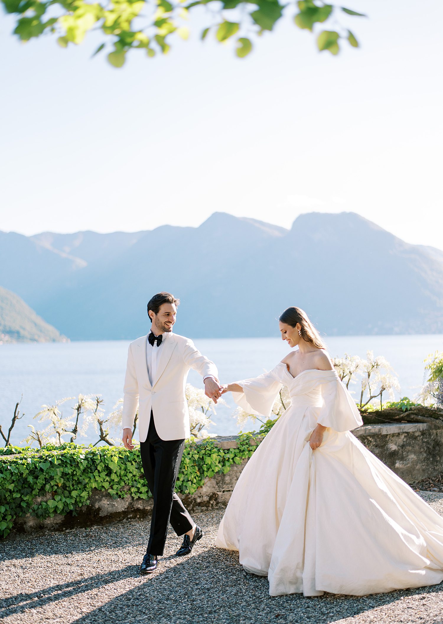 bride and groom hold hands walking in front f Lake Como