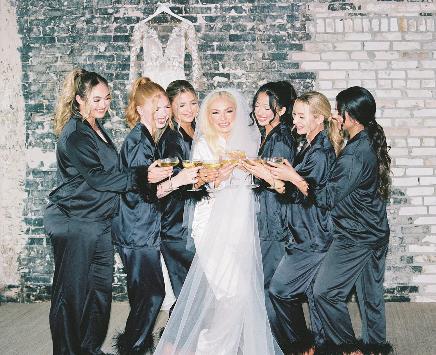 bridesmaids toast bride in front of blue brick wall on morning of wedding