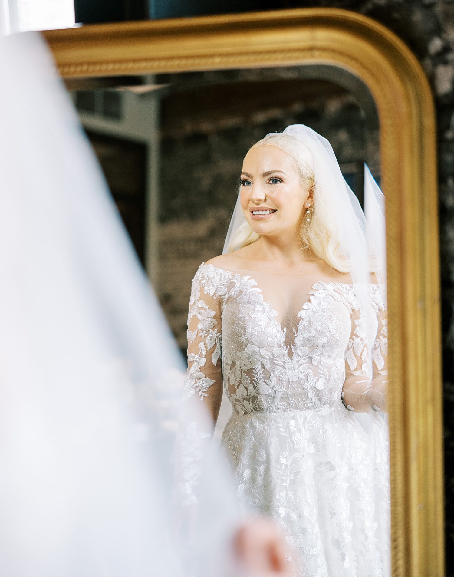  bride smiles at herself in the mirror with veil around her shoulders 