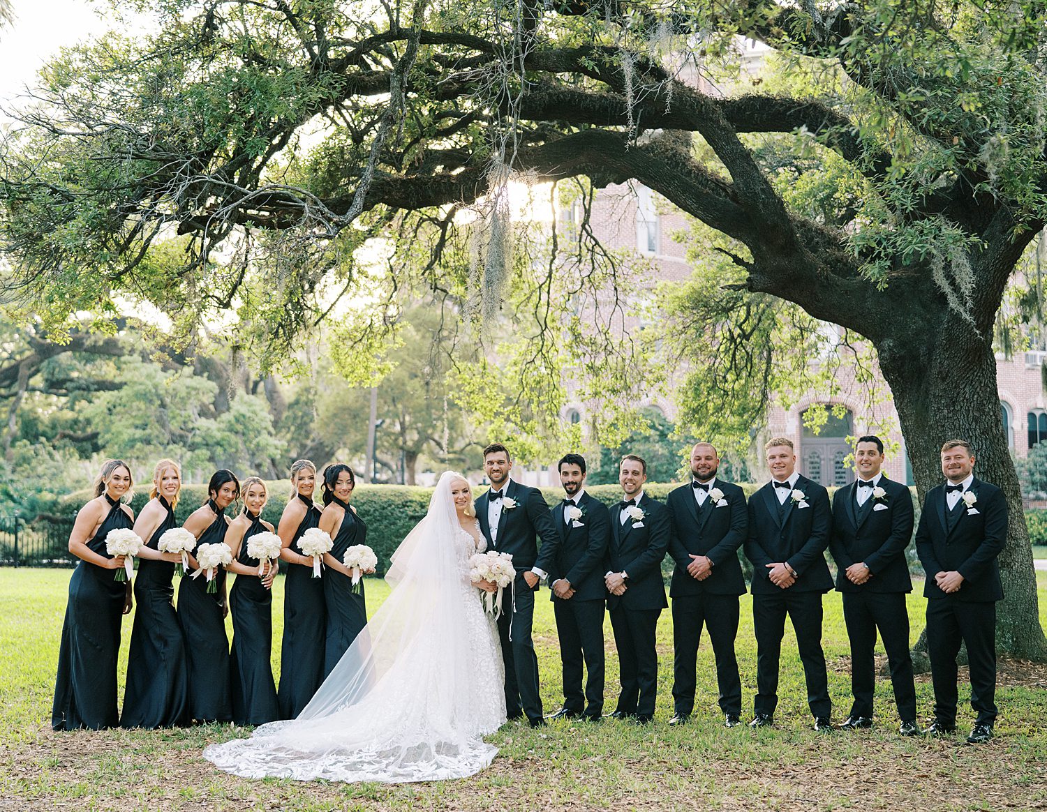 bride and groom stands with wedding party in all black under tree at the Oxford Exchange