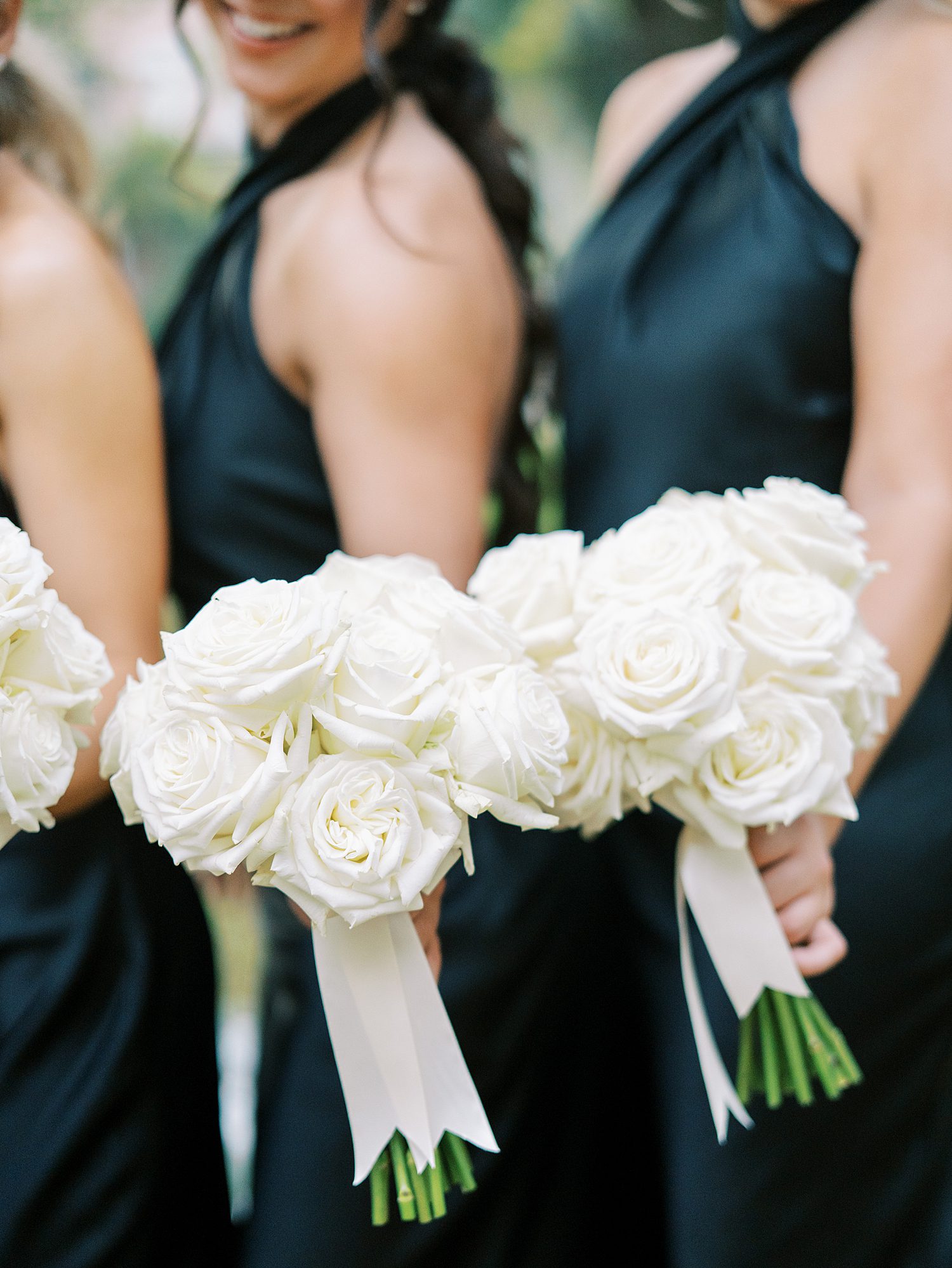 bridesmaids in black gowns hold bouquets of ivory roses with white ribbons
