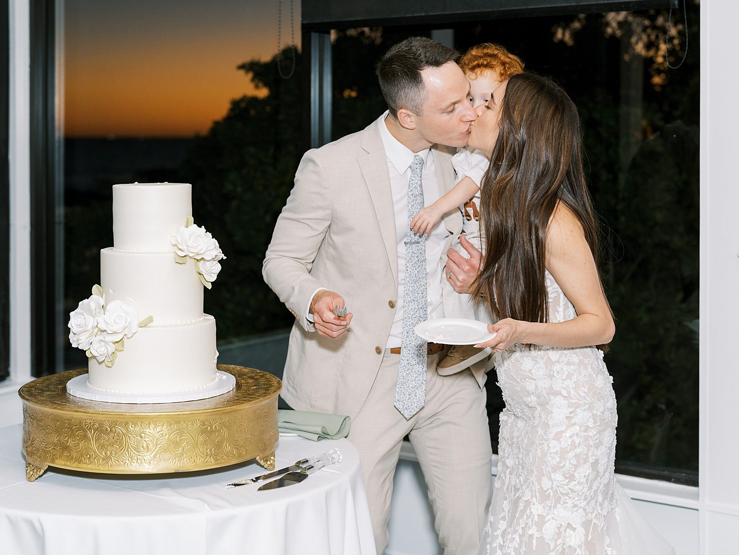 bride and groom cut wedding cake and kiss son on their hip