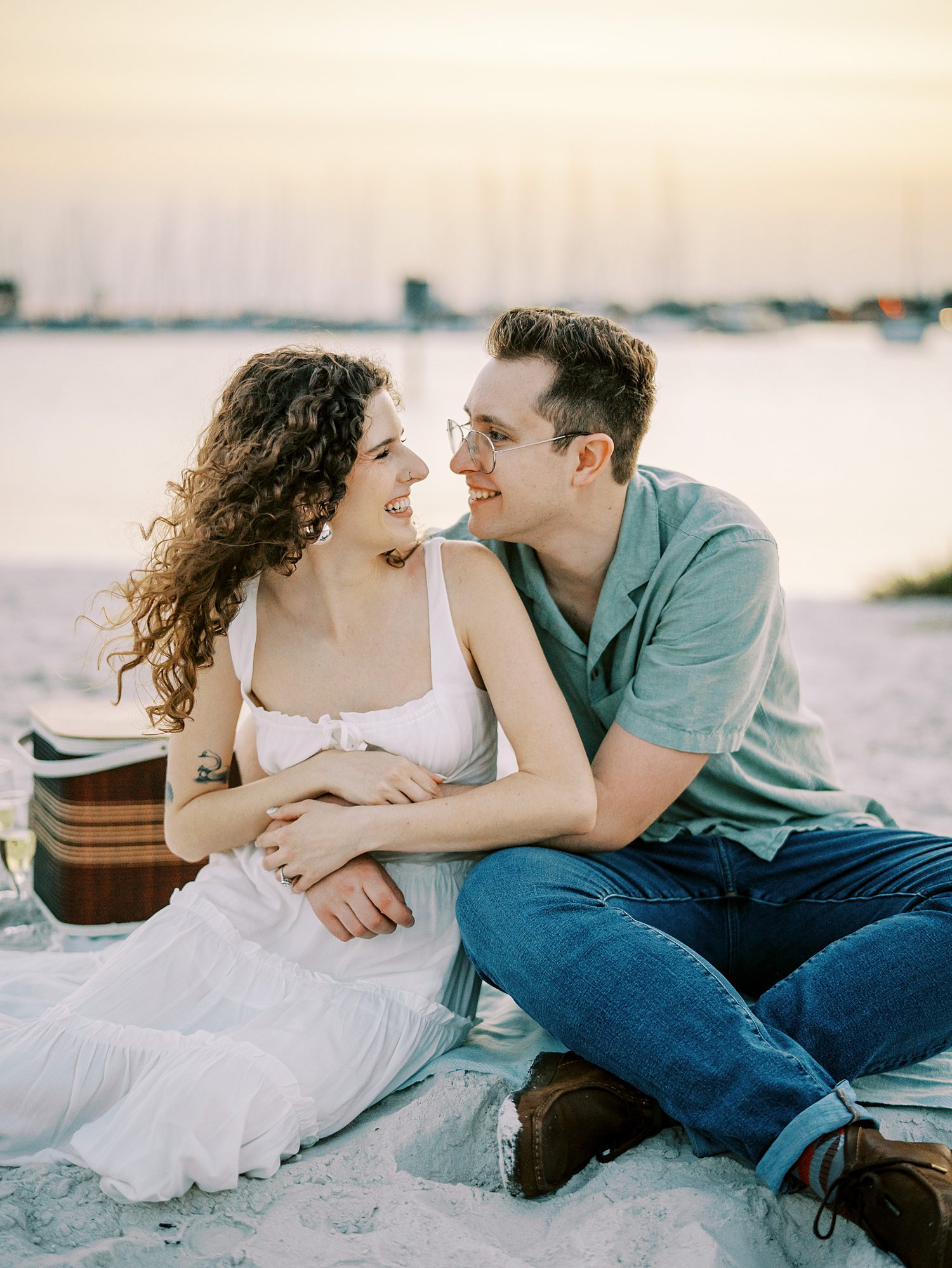 man in green polo hugs woman in white dress around waist sitting on beach