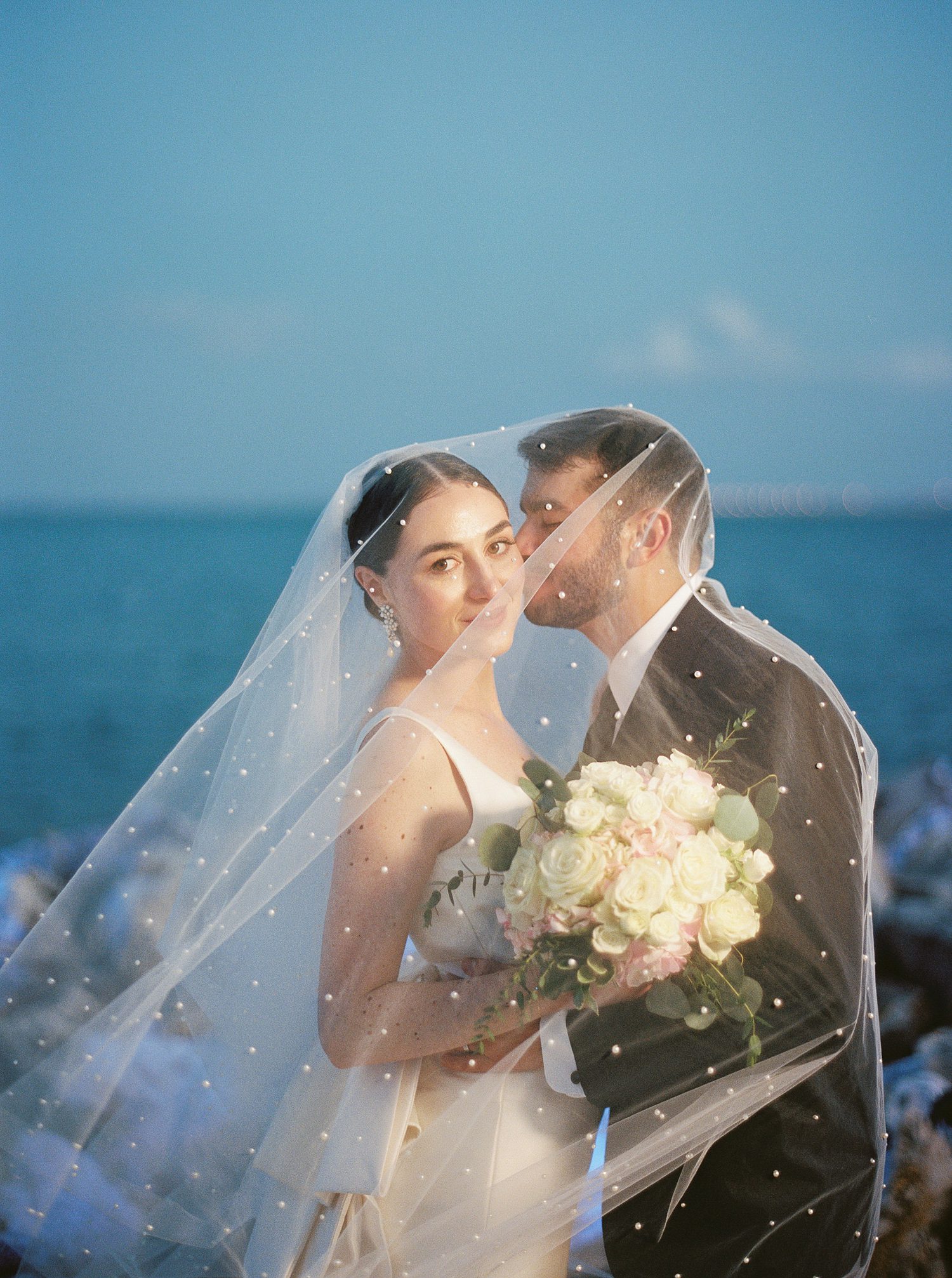 groom kisses bride's cheek under veil on rocky Tampa FL beach